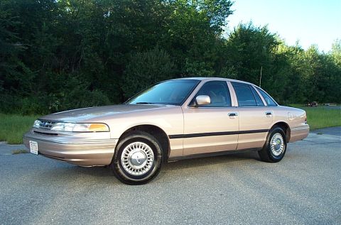 2001 Ford Crown Victoria - A tan 2001 Ford Crown Victoria sedan is parked on an asphalt surface with trees in the background.