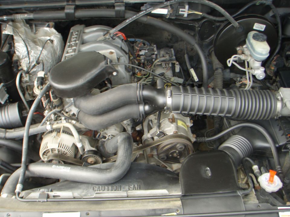 Engine bay with hoses, belts, and engine components visible. Text on fan shroud reads 'CAUTION - FAN'