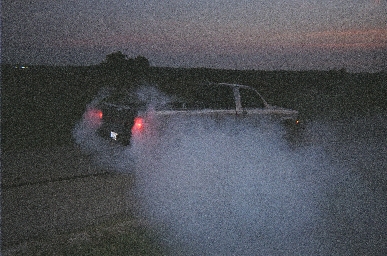 1988 Ford LTD Crown Victoria - Grainy dusk shot of a dark sedan doing a burnout on a rural road, engulfed in a cloud of white tire smoke with its red taillights and brake…
