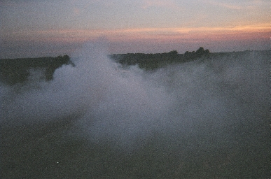 1988 Ford LTD Crown Victoria - A dense bank of white tire smoke rolling across a dark rural road at dusk, with a pink and orange sunset over a tree line on the horizon. N…