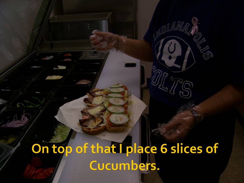 Sandwich with meat, cheese, and cucumber slices on a white paper, with a person in an Indianapolis Colts shirt preparing it