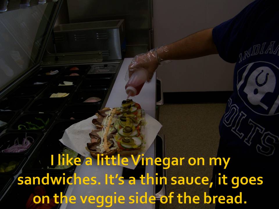 person adding sauce to a large sandwich at a food prep station