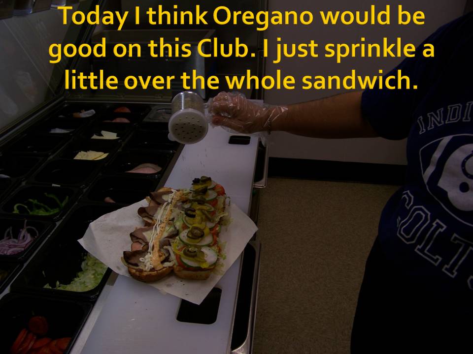 Sandwich being seasoned with oregano over a white paper wrapper on a counter with salad bar ingredients in the background