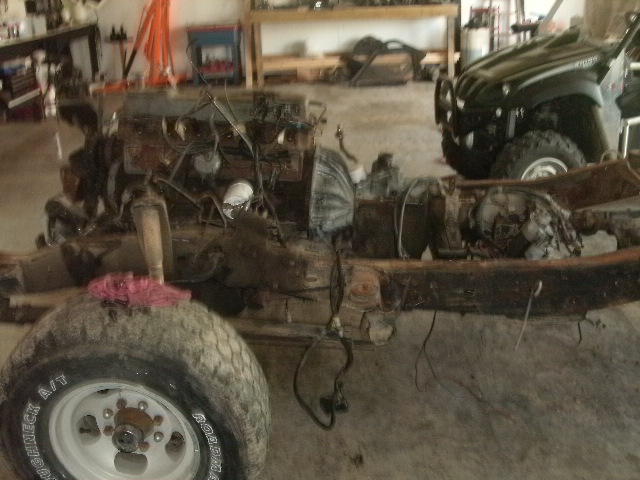 Bare truck frame with engine and transmission exposed in a garage, white wheels with Roughneck A/T tires
