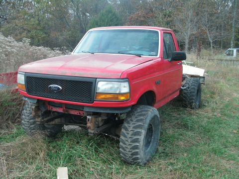 1995 Ford F-150 - Front three-quarter view of a lifted red Ford F-150 regular cab sitting in tall grass with its bed removed, exposing the bare rear frame. I…