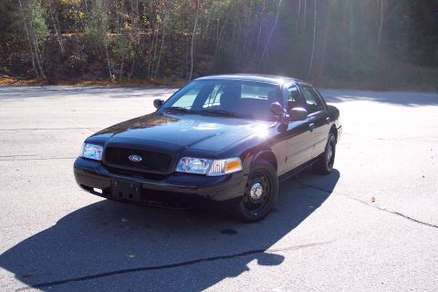 2001 Ford Crown Victoria - A black 2001 Ford Crown Victoria police sedan is parked on asphalt with trees in the background.
