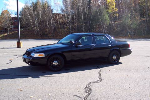 2001 Ford Crown Victoria - Black 2001 Ford Crown Victoria police car parked in a lot with trees in the background.