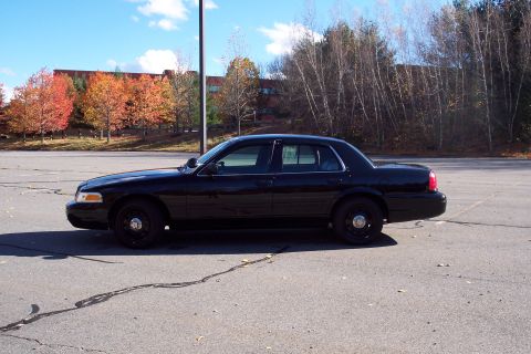 2001 Ford Crown Victoria - A black sedan is parked in a lot during autumn, with colorful trees and a building visible.