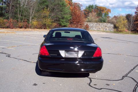 2001 Ford Crown Victoria - A black sedan is parked in an empty lot with trees and a stone wall behind it.