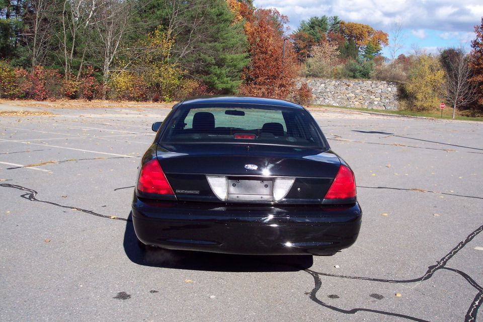 Rear view of a black sedan parked in a lot. Trees and a stone wall are in the background