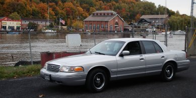 2000 Ford Crown Victoria - A silver 2000 Ford Crown Victoria is parked near a body of water with buildings and trees visible.