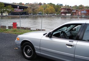 2000 Ford Crown Victoria - A silver 2000 Ford Crown Victoria is parked near a body of water with industrial buildings in the background.