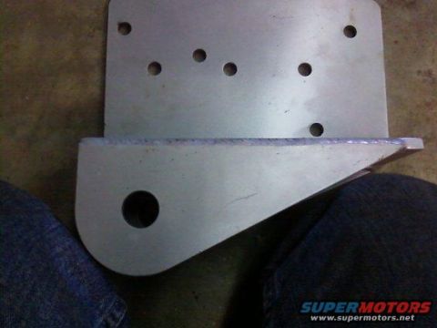 1995 Ford Bronco - A metal bracket with several drilled holes and a prominent circular opening is shown, with a portion of jeans visible at the bottom.