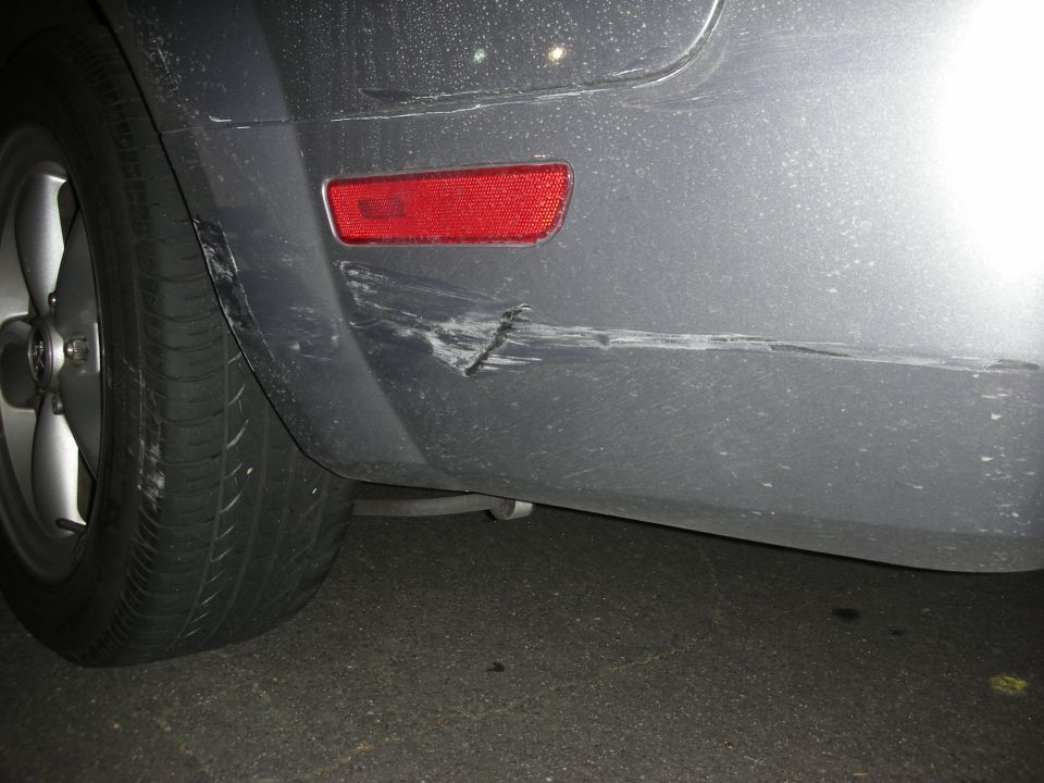Gray car's rear bumper with a deep scratch and scuff marks near a red reflector and wheel