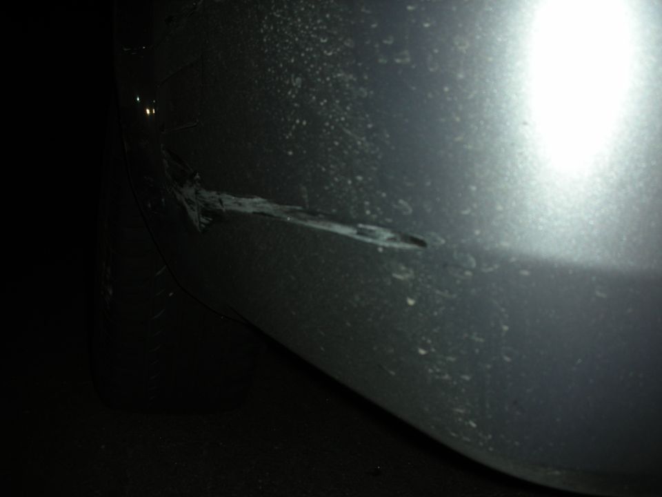 Close-up of a scraped and dented silver car bumper with some dirt on it