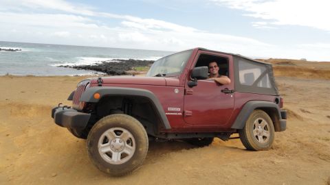 1989 Ford Bronco - A red Jeep Wrangler is parked on a sandy beach with the ocean in the background.