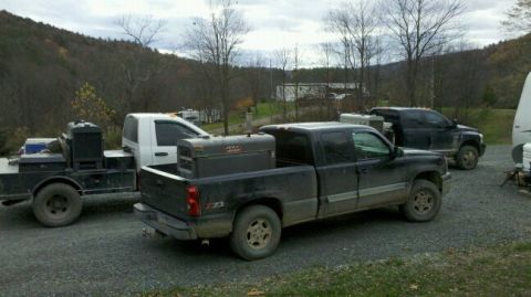 2003 Chevrolet Silverado 1500 - Two pickup trucks are parked outdoors, one with a welding machine in its bed, near a trailer and a wooded hillside.