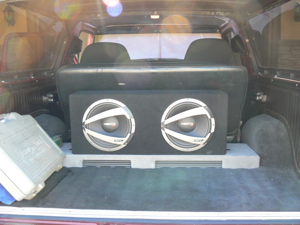 Interior cargo area of a vehicle showing a dual subwoofer box and a grey storage container