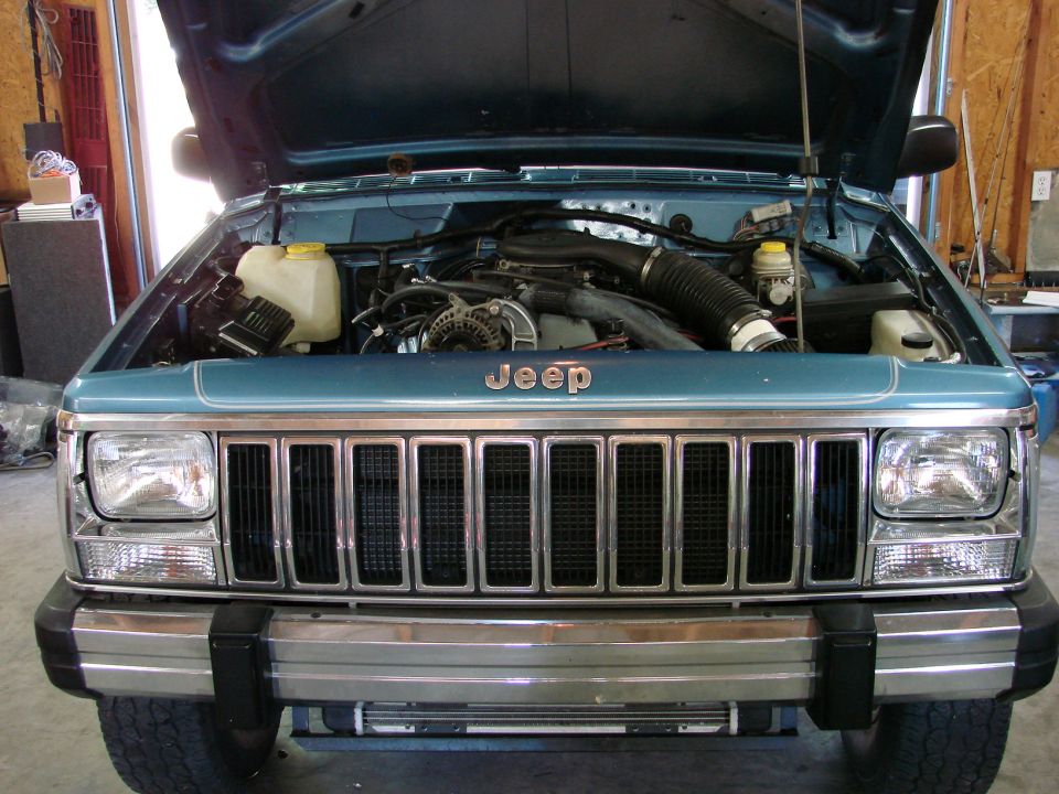 Front view of a teal Jeep Cherokee with hood open showing the engine and grille. The Jeep logo is visible above the grille