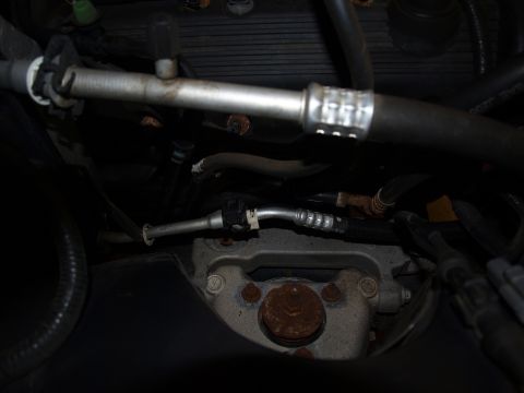 1998 Ford Crown Victoria - Close-up down into a dim engine bay showing steel fuel or heater lines with crimped quick-connect couplings running across a ribbed black v…