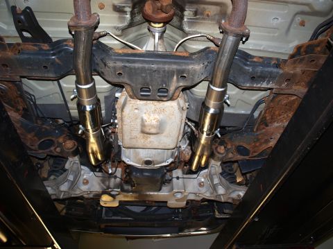 1998 Ford Crown Victoria - Underside view of a car on a lift showing new stainless dual exhaust mid-pipes clamped to rusty factory manifold downpipes, with heat-tinte…