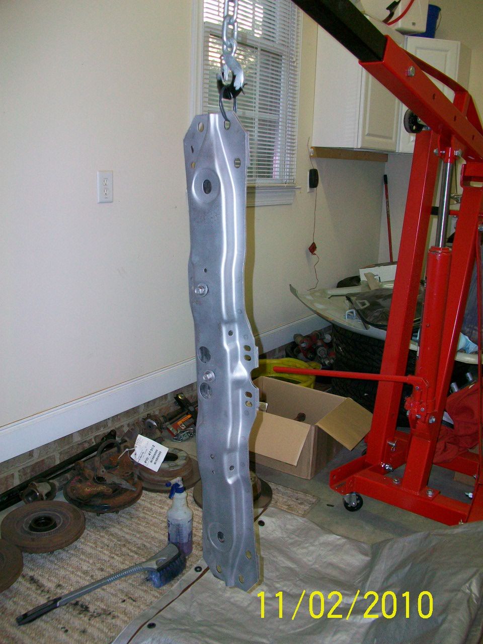 metal car frame piece hanging from engine hoist, with brake rotors and cleaning supplies on floor