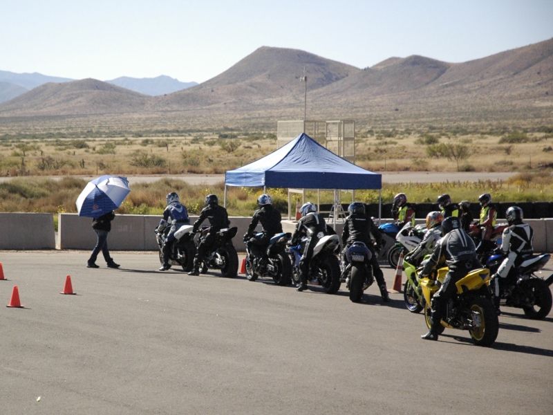 Motorcycles and riders gathered on a track with a blue tent and mountains in the background