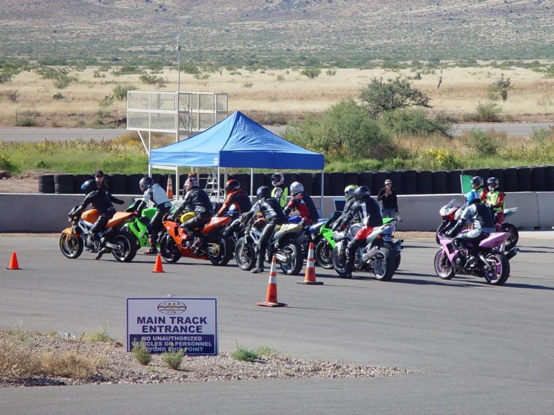 Group of motorcycles lined up on a race track with riders in gear. A blue tent is set up trackside