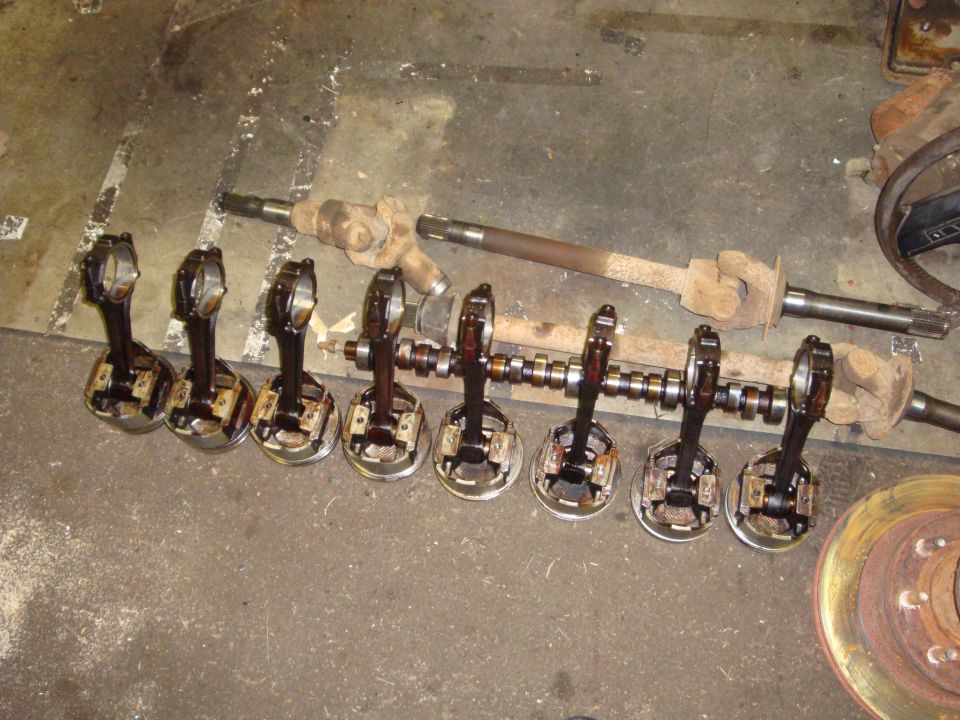 Eight pistons with connecting rods attached to a crankshaft, with driveshafts and a brake rotor in the background