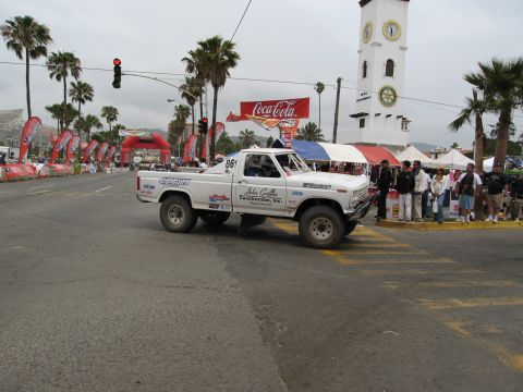 1986 Ford Bronco - Off-road pickup truck competes in a race, passing under an archway with spectators watching.