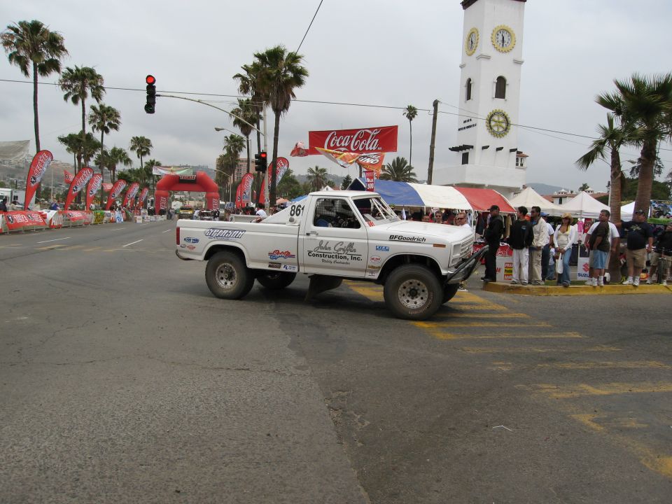 White off-road pickup truck with racing decals, number 861, driving on a street with palm trees and banners
