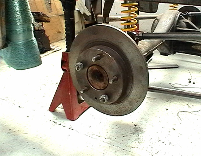 Car brake rotor assembly on a red jack stand, with yellow coil spring and suspension parts visible