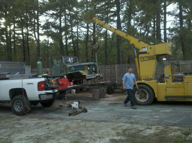 Green military truck on a trailer, being lifted by a yellow Pettibone crane. A white pickup truck is parked nearby