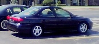 1999 Ford Taurus - A dark 1999 Ford Taurus sedan is seen from the side, parked next to another vehicle.