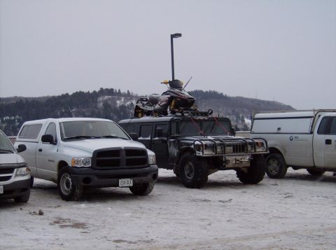 1981 Ford Bronco - A Humvee is parked in the snow with a snowmobile loaded onto its roof rack, alongside other vehicles.