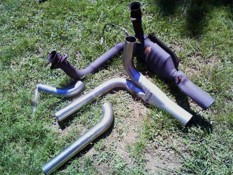 1990 Ford Bronco - Old and new exhaust parts laid out side by side on sunlit grass: a rusty factory Y-pipe with catalytic converter and an oxygen sensor still…