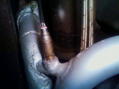1990 Ford Bronco - Close-up of a painted-silver exhaust pipe with an oxygen sensor threaded into a welded bung, set against a dark engine or transmission and…