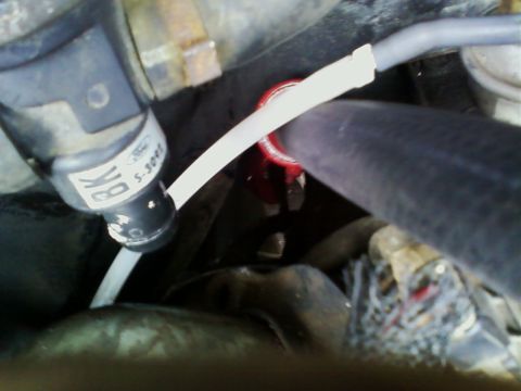 1990 Ford Bronco - Blurry close-up deep in an engine bay of a black vacuum solenoid/valve marked with a Ford oval and part number, with a white plastic vacuum…