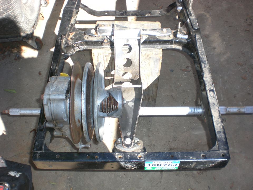 Bare black frame with rear axle, transaxle and pulleys on concrete; small California plate 18K76J attached.