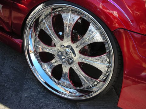 1998 Ford Crown Victoria - polished chrome multi-spoke wheel with ornately engraved spokes mounted on a red car, low-profile tire visible.
