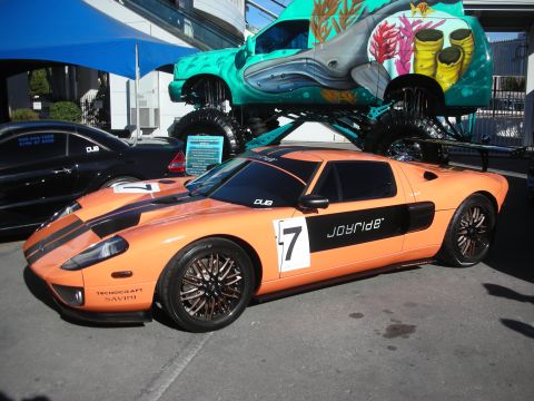 1998 Ford Crown Victoria - orange Ford GT with black stripes and 'Joyride' number-7 graphics parked beside a sea-themed monster truck.