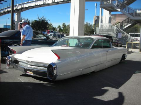 1998 Ford Crown Victoria - white finned Cadillac coupe sits lowered, rear three-quarter view, under a pedestrian bridge at an outdoor event.