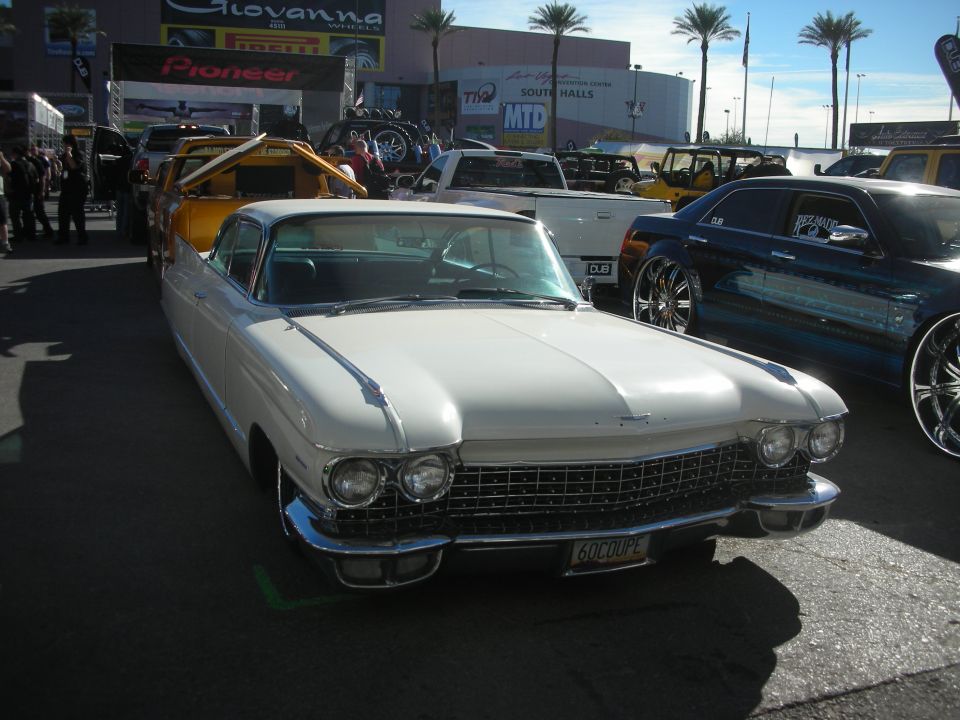 White 1960 Cadillac at an outdoor car show with palm trees and vendor tents