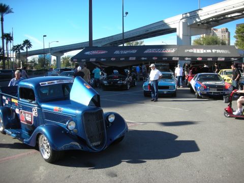 1998 Ford Crown Victoria - blue vintage hot-rod pickup with race numbers parked among show cars beneath a highway overpass.