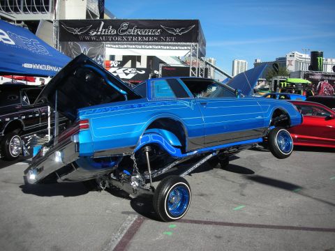 1998 Ford Crown Victoria - custom blue lowrider coupe sits tipped up on its hydraulic suspension and chrome wire wheels at an outdoor car show.