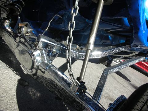 1998 Ford Crown Victoria - Close-up of an engraved chrome rear axle, driveshaft and hydraulic cylinder on a blue lowrider parked on asphalt.