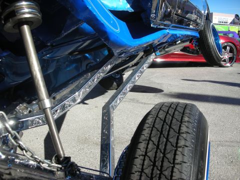 1998 Ford Crown Victoria - Detail of polished engraved suspension links and a hydraulic ram under a blue lowrider, with a tire and red car nearby.