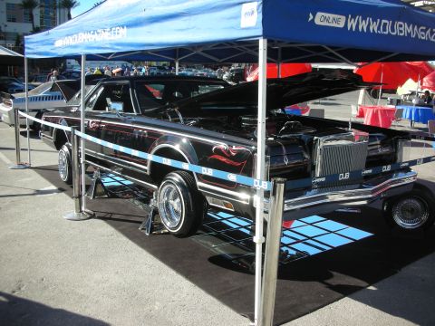 1998 Ford Crown Victoria - black custom coupe with red and blue pinstriping sits raised on a glass display stand beneath a branded show tent.