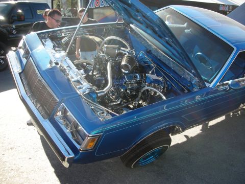 1998 Ford Crown Victoria - blue custom car displays its open hood and heavily chromed, dressed engine bay while spectators look on at a show.