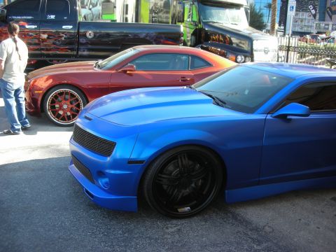 1998 Ford Crown Victoria - matte-blue muscle coupe on black wheels sits beside an orange one with bronze wheels at an outdoor car show.
